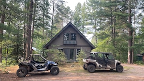 Deux véhicules tout-terrain sont stationnés devant une cabane entourée d’arbres.