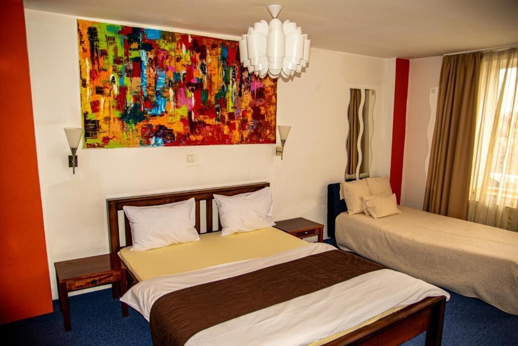 Hotelkamer met een groot bed, een kleiner bed, een kleurrijk abstract schilderij aan de muur, een witte kroonluchter en een raam met gordijnen.