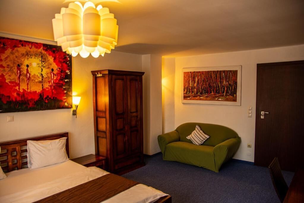 Une chambre d’hôtel avec un lit, un fauteuil vert, une armoire en bois, un tableau au mur et un luminaire au plafond.