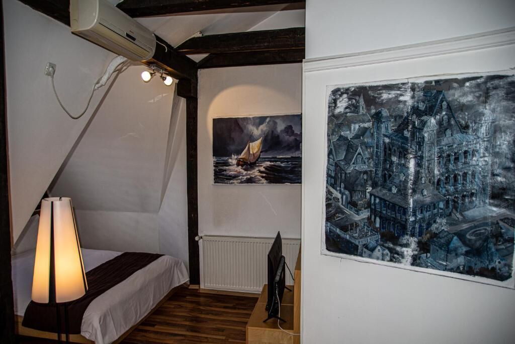 A bedroom with a bed, a desk, a lamp, a painting of a ship on the sea, and a large framed artwork of a cityscape.