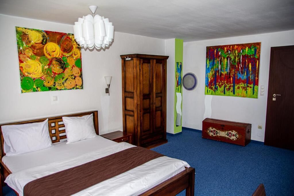 A bedroom with a bed, a wardrobe, two paintings, and a small chest of drawers.