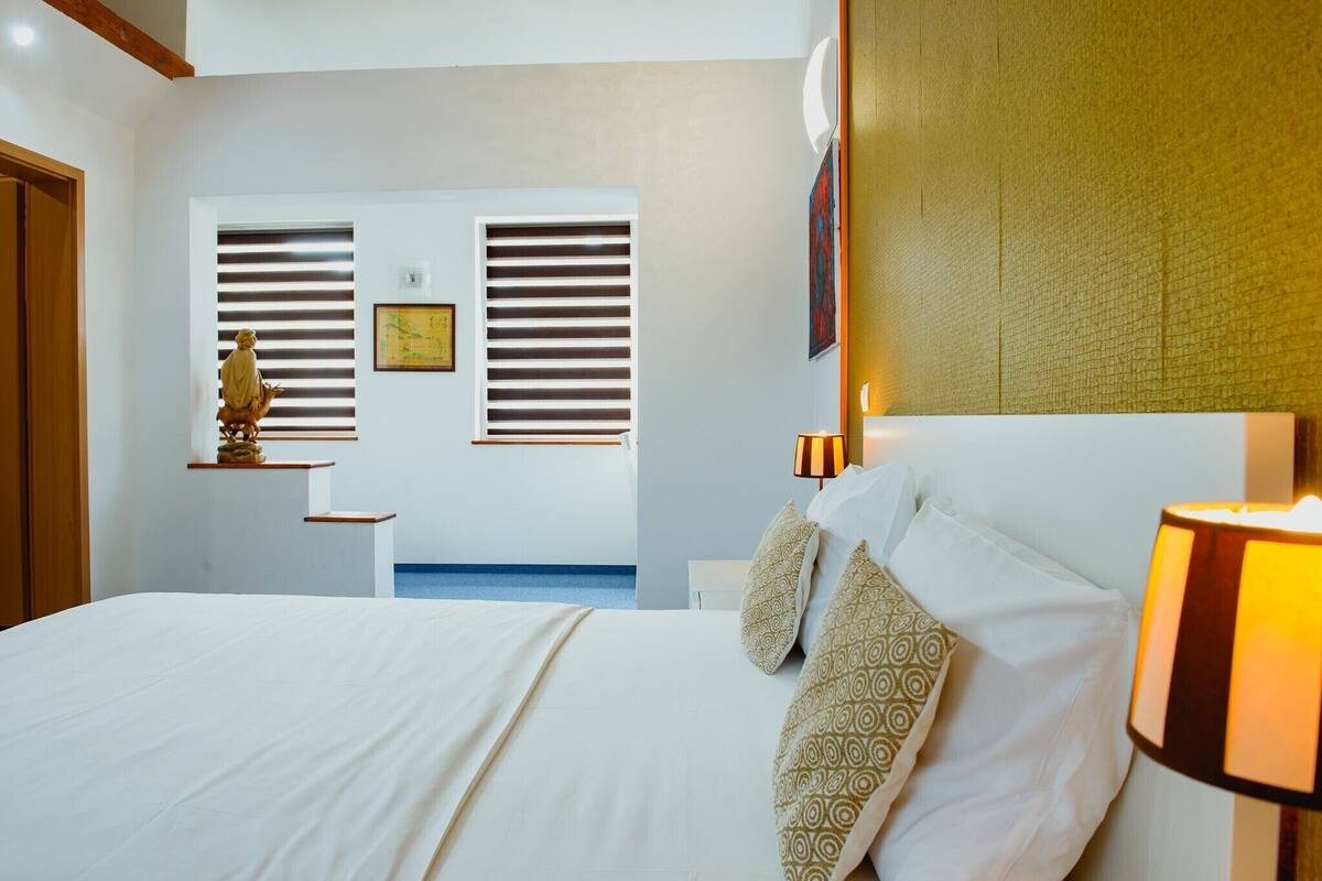 A hotel room with a large bed, a nightstand with a lamp, a statue on a shelf, and a window with blinds.