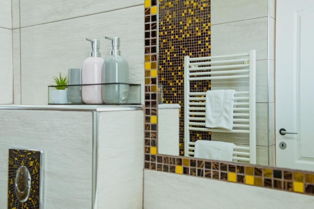 Un baño con un lavamanos blanco, dos botellas de jabón líquido, un recipiente de vidrio con una planta y un perchero con una toalla blanca.