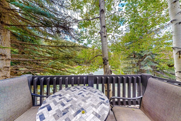 A round table with a herringbone pattern tabletop, surrounded by cushioned chairs with a woven seat design, set against a backdrop of a wooden fence and dense green foliage.
