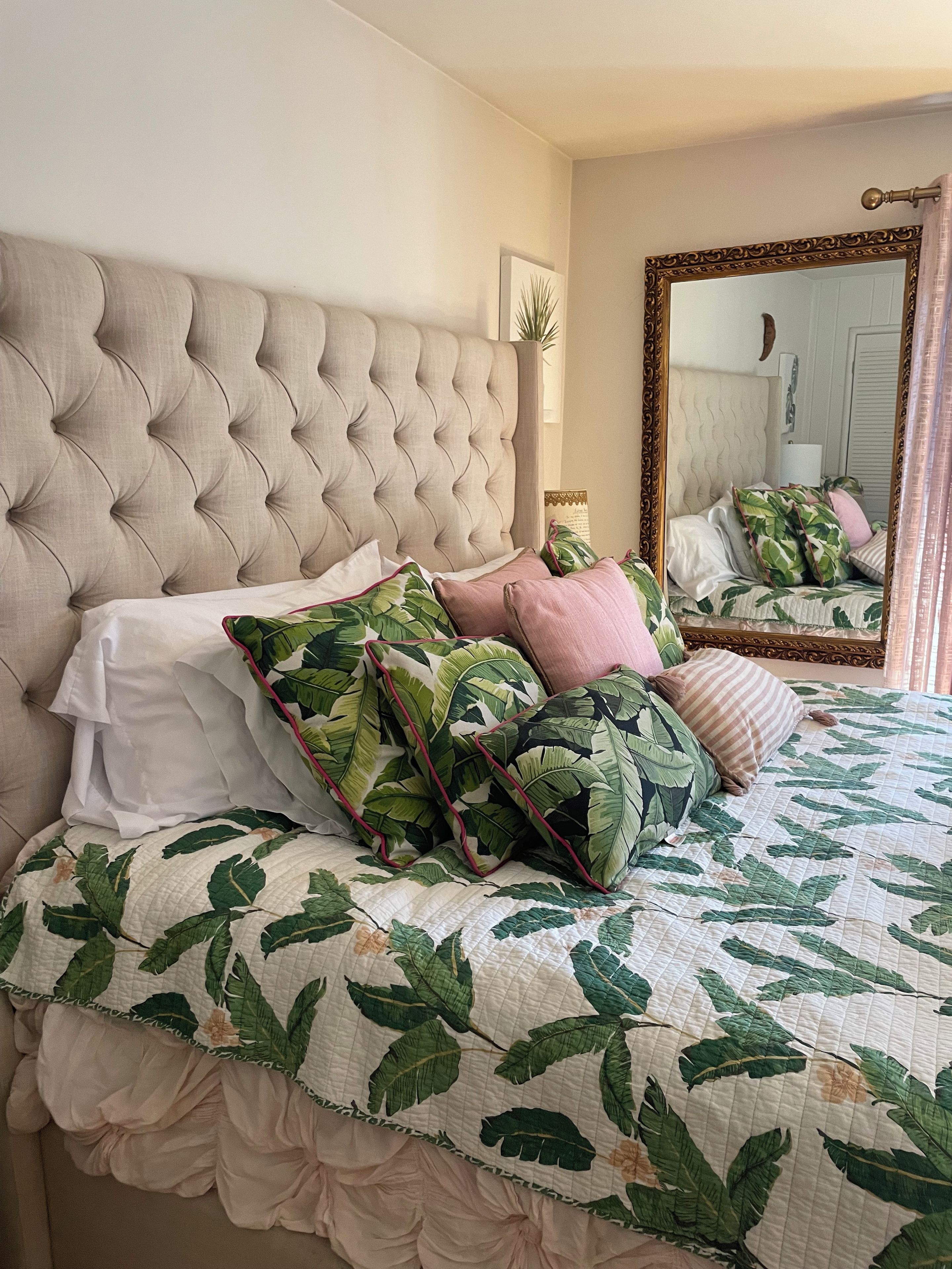 Un dormitorio con cabecera capitonada, cama con colcha estampada de hojas y espejo con marco decorativo.