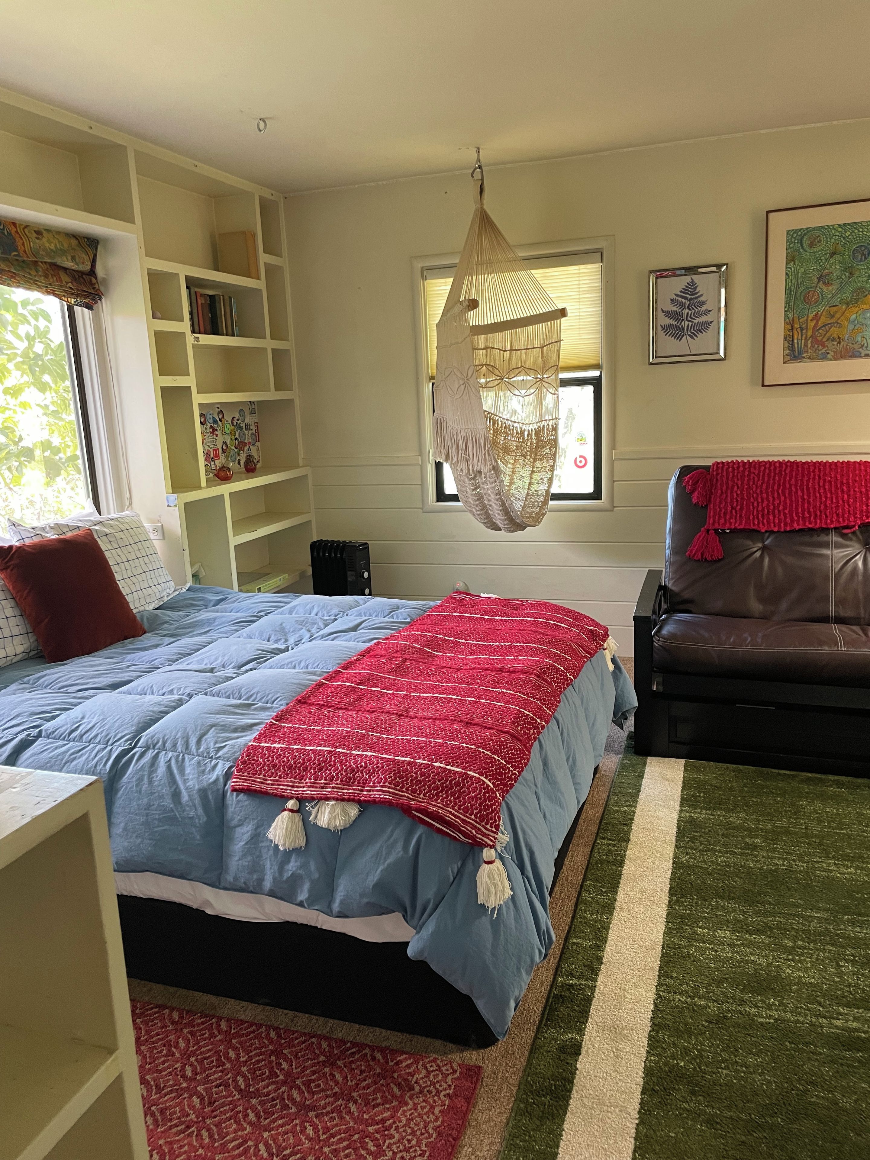 A bedroom with a bed, a hanging macrame chair, a bookshelf, a window with blinds, and a couch.