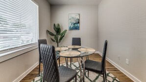 Een eetgedeelte met een glazen tafel, vier stoelen, een potplant en een schilderij aan de muur.