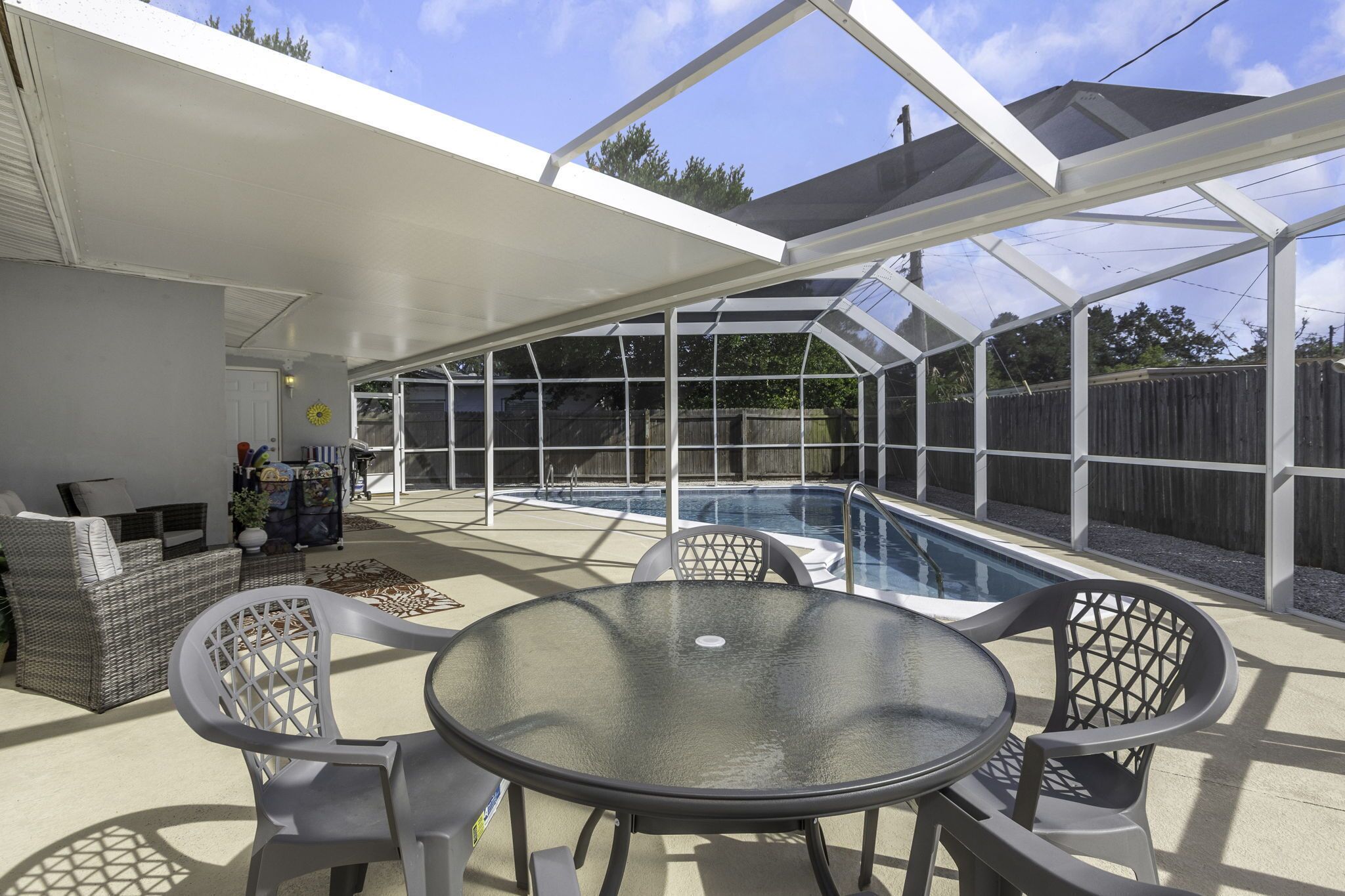 A covered patio with a glass enclosure, a pool, outdoor furniture, and a wooden fence in the background.