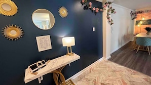 Un mur bleu foncé orné de miroirs décoratifs en forme de soleil, un miroir rond et un petit tableau encadré. On y voit une console blanche avec une lampe et un objet décoratif. Le sol présente un motif en chevrons et une chaise est visible.