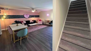 Un salon moderne avec un canapé, une table basse et un coin repas. Un escalier est visible sur la droite.