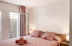 A hotel room with a bed, two bedside lamps, a window with blinds, and a door with a curtain.
