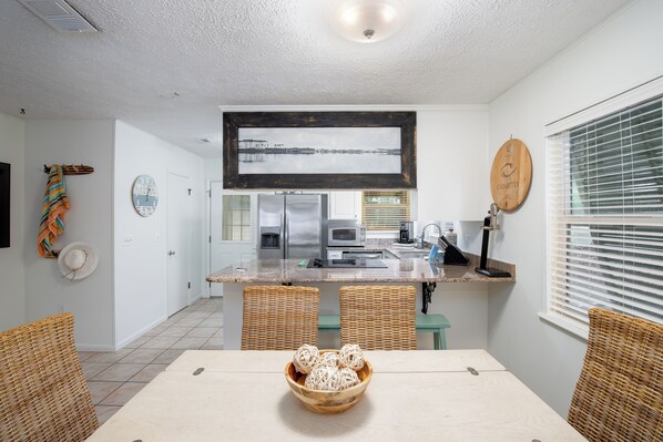 A modern kitchen with a central island, wicker chairs, and a dining table with a bowl of decorative items. - Steps to the Beach - On 30A - Walk to Seaside (Santa Rosa Beach)
