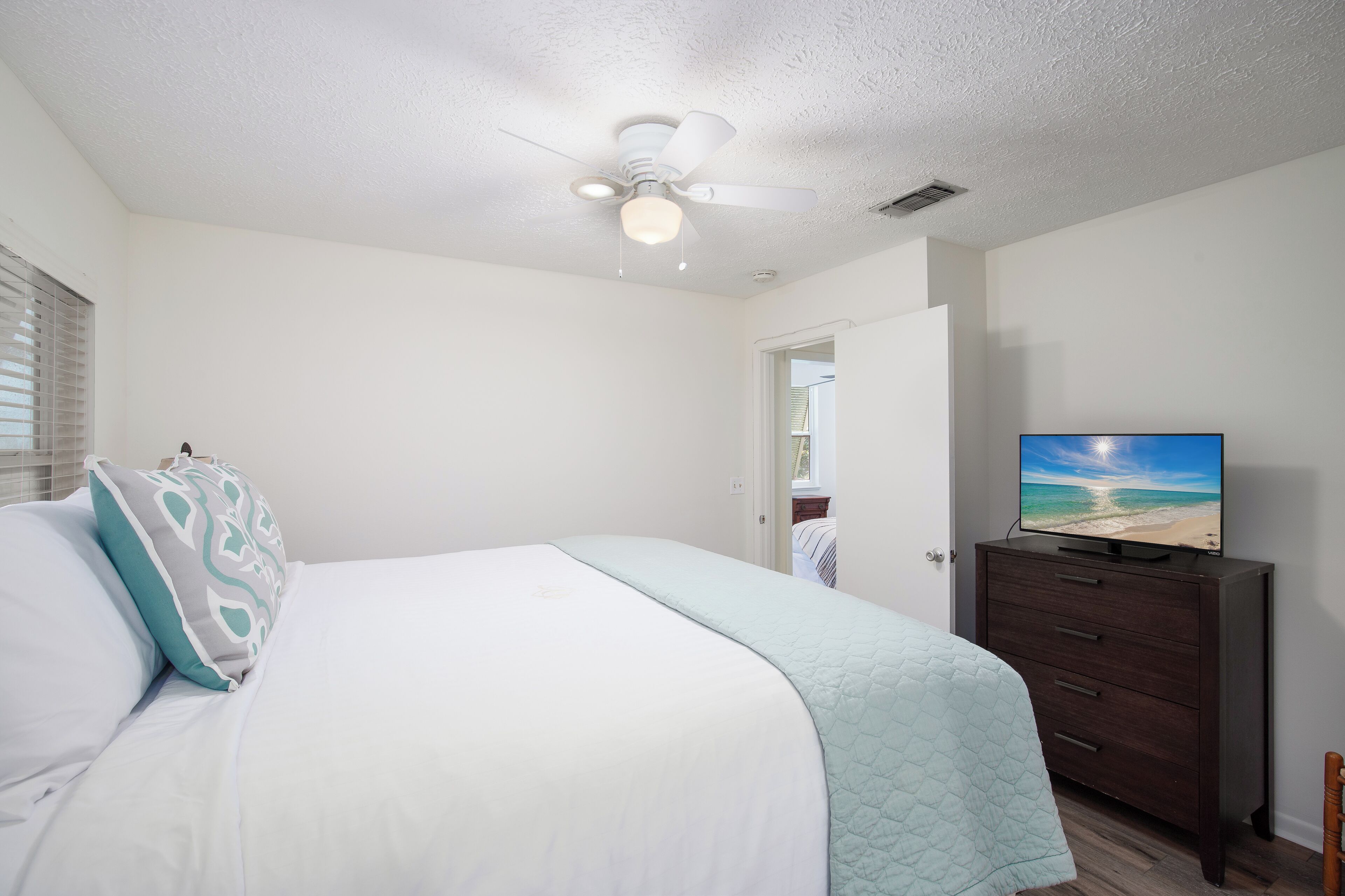 A bedroom with a bed, a ceiling fan, a dresser, and a TV displaying a beach scene.