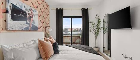 A bedroom with a bed, a TV, a window with curtains, a plant, and a balcony view.