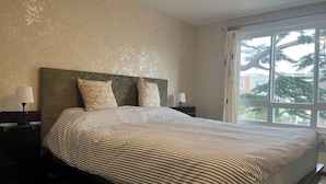 A bedroom with a large bed, two bedside tables with lamps, a window with a view of trees, and patterned wallpaper.