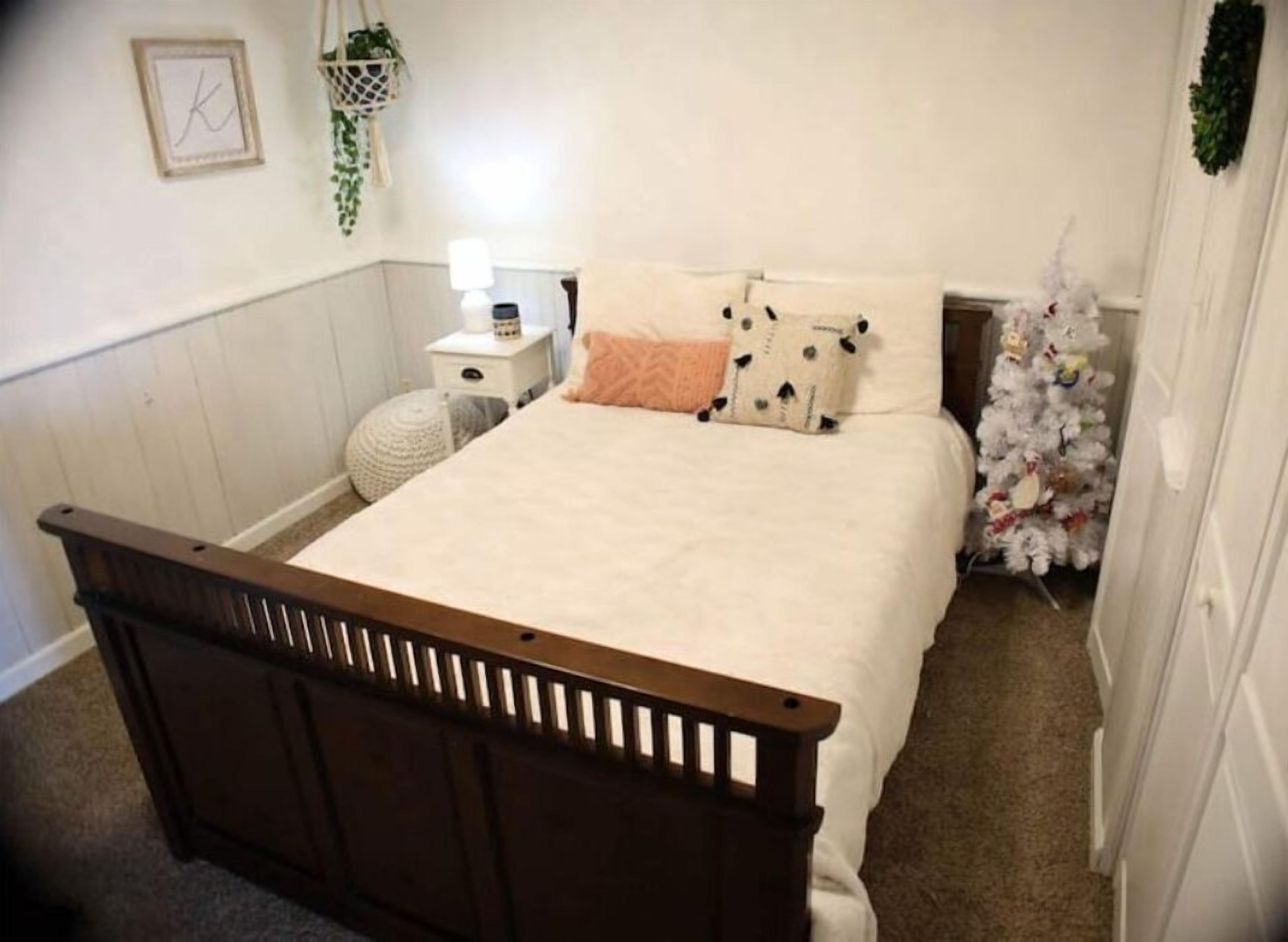 A bedroom with a wooden bed frame, white bedding, a small bedside table with a lamp, and a white Christmas tree.