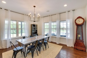 Une salle à manger avec une grande table, des chaises, un lustre, une horloge et de grandes fenêtres avec des rideaux.