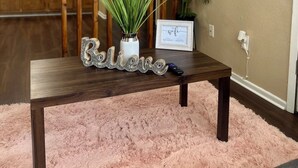 A living room with a wooden TV stand, a pink fluffy rug, a 'Believe' sign, a framed photo, and a potted plant. - Eagle Stay 7 Condo-Eagle Pass (Eagle Pass)