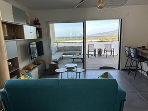 Ett modernt vardagsrum med en turkosfärgad soffa, en tv och en balkong med utsikt över havet.