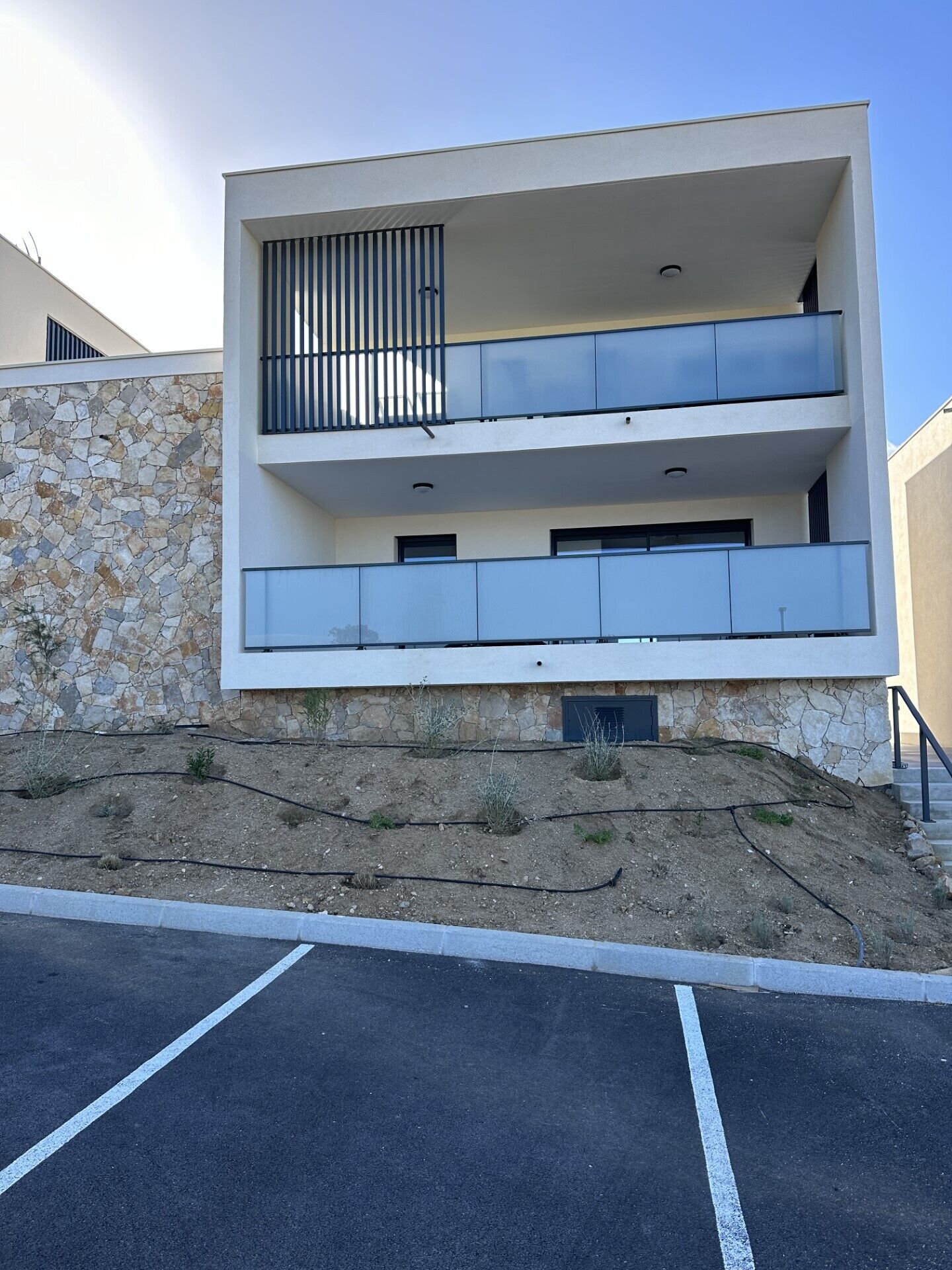 Un edificio moderno a due piani con facciata in pietra, balconi e un parcheggio antistante.