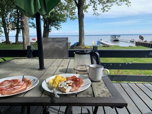 A wooden table with breakfast plates, a mug, and a kettle on a deck overlooking a lake.
