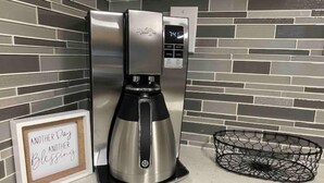 Une cafetière en acier inoxydable dotée d’un écran numérique, d’un panneau encadré portant l’inscription « Encore un jour, encore une bénédiction », et d’un panier en fil de fer noir posé sur un comptoir dont le dos est recouvert de carreaux.