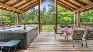 A covered outdoor deck with a hot tub, dining table, and chairs, overlooking a grassy area and trees. - Blue Water Dunes Cottage (Tiny)