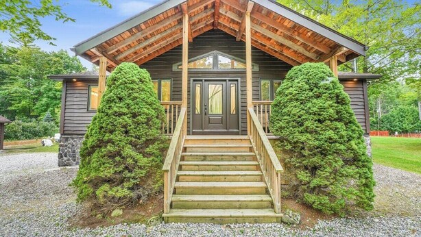 A log cabin with a wooden staircase leading to a front door, surrounded by two large evergreen trees. - Blue Water Dunes Cottage (Tiny)