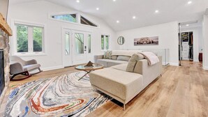 A spacious living room with a large sectional sofa, a coffee table, and a colorful rug. There are large windows providing natural light and a view of greenery outside. - Blue Water Dunes Cottage (Tiny)