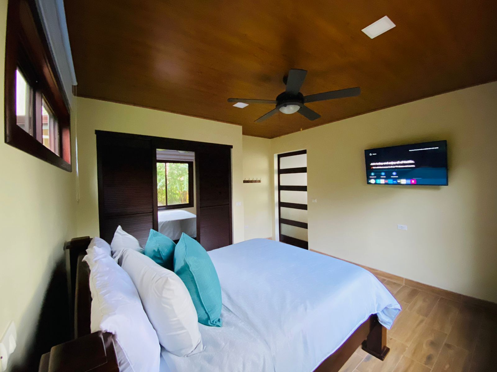 Un dormitorio con cama, almohadas, ventilador de techo, televisión y ventana.