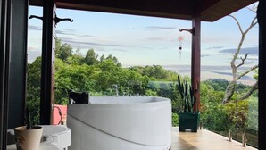 A modern bathroom with a freestanding bathtub, wooden flooring, and a view of lush greenery.