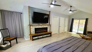 A modern bedroom with a large bed, a fireplace, a TV mounted on a wall, a ceiling fan, and a view of the outdoors through a window. - Mountain Ridge Retreat - Private & Peaceful Getaway. Family & Pet Friendly (West Park)