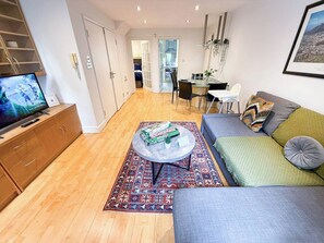 Ein modernes Wohnzimmer mit einer Couch, einem Couchtisch mit Gesellschaftsspielen, einem Fernseher und einem Essbereich im Hintergrund.