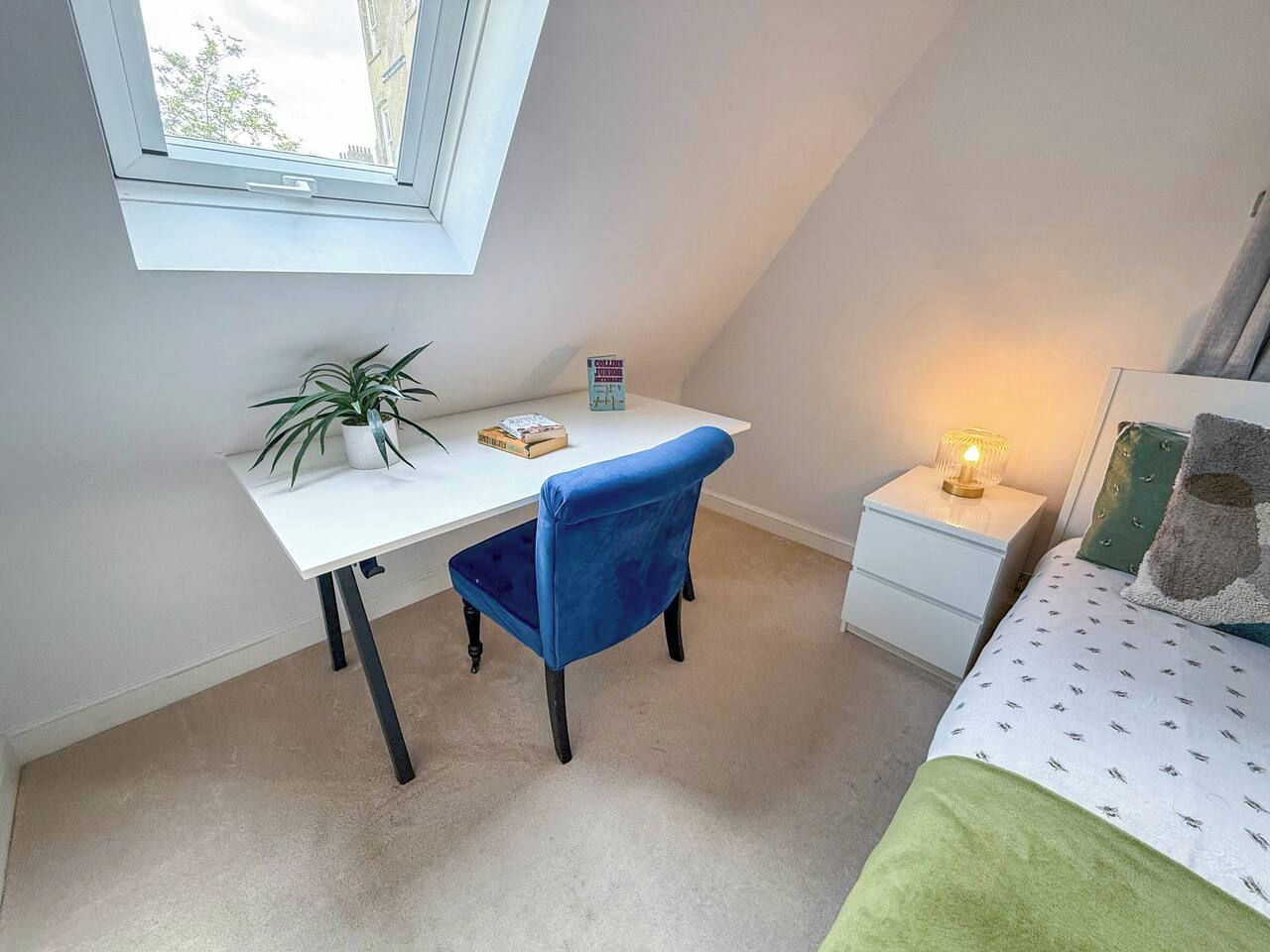 A bedroom with a bed, a desk, a chair, a nightstand, a lamp, and a window.
