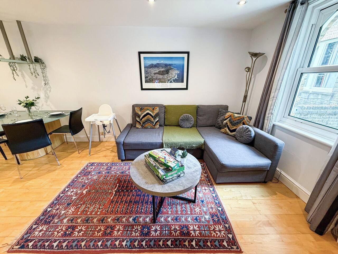 Een woonkamer met een bank, een salontafel, een kamerbreed tapijt met een patroon, een eethoek met stoelen, en een ingelijst schilderij aan de muur.
