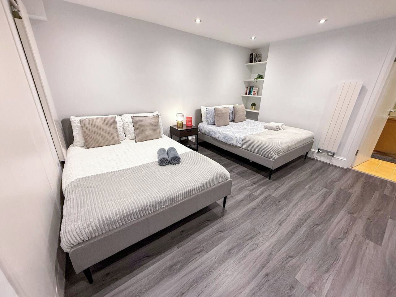 A modern bedroom with two beds, a bedside table with a lamp, and a built-in shelf unit.