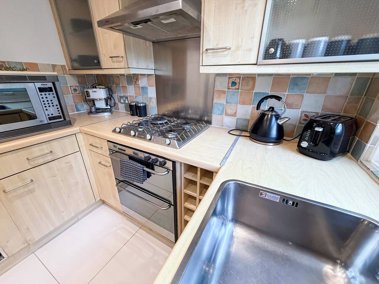 A compact kitchen with a gas stove, oven, microwave, toaster, kettle, and a stainless steel sink.