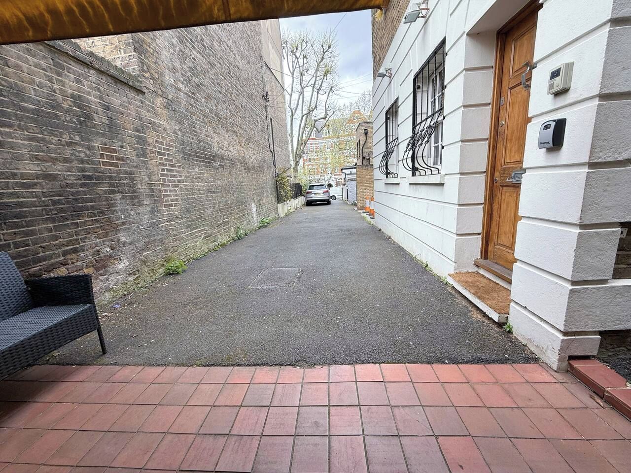 A narrow alleyway with a brick bench on the left, a white building with a wooden door on the right, and a car parked in the distance.