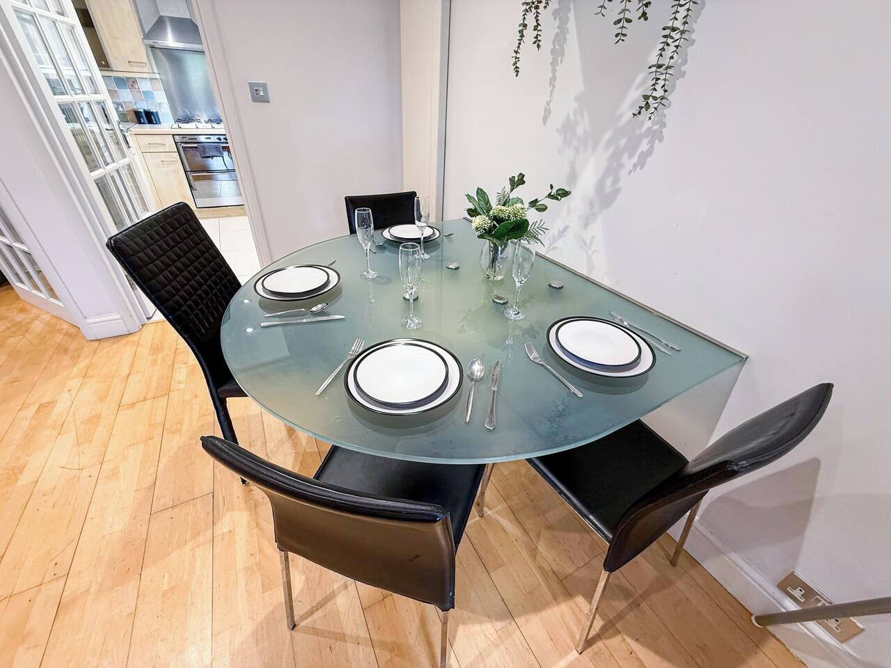 A round glass table with four chairs, a vase with greenery, and a kitchen in the background.