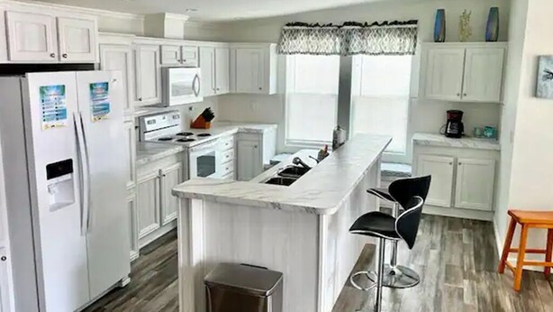 Una cocina moderna con armarios blancos, una isla de mármol, taburetes negros y suelo de madera.