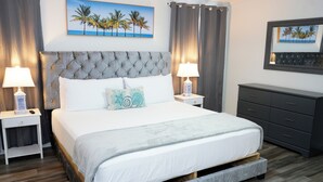 Un dormitorio ordenado con tablero de cama capitonado, una cama grande con ropa de cama blanca, dos mesitas de noche con lámparas, un tocador y dos cuadros en la pared.