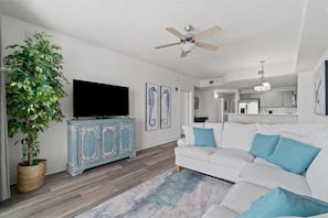 Un salon avec un canapé blanc, un ventilateur de plafond, un téléviseur à écran plat, un meuble bleu et une grande plante.