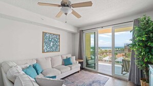 A modern living room with a white sofa, blue cushions, a ceiling fan, and a large sliding glass door leading to a balcony with a view of the ocean.