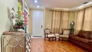 Una sala de estar con piso de madera, un sofá de cuero marrón, dos sillones, una mesita y un mueble de consola decorado con plantas.