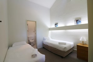 Two single beds with white linens, a nightstand, and a dresser in a compact room with a window and a door to a bathroom.