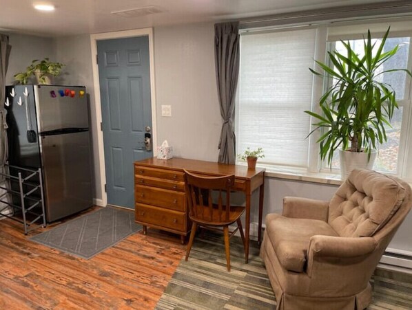 A compact living space with a kitchenette, a wooden dresser, a cushioned armchair, and a potted plant by a window. - The Fox Den (Ames)