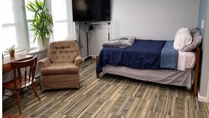A compact room with a bed, a chair, a TV, and a window with a plant. - The Fox Den (Ames)