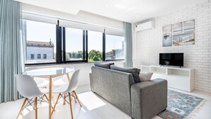 A modern living room with a grey sofa, white chairs, a TV on a white cabinet, and large windows offering a city view. - VIVA  Marquês Studio (Porto)