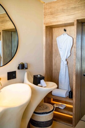 A modern bathroom with a large mirror, two sinks, a robe hanging on a wooden door, and a small shelf with toiletries.