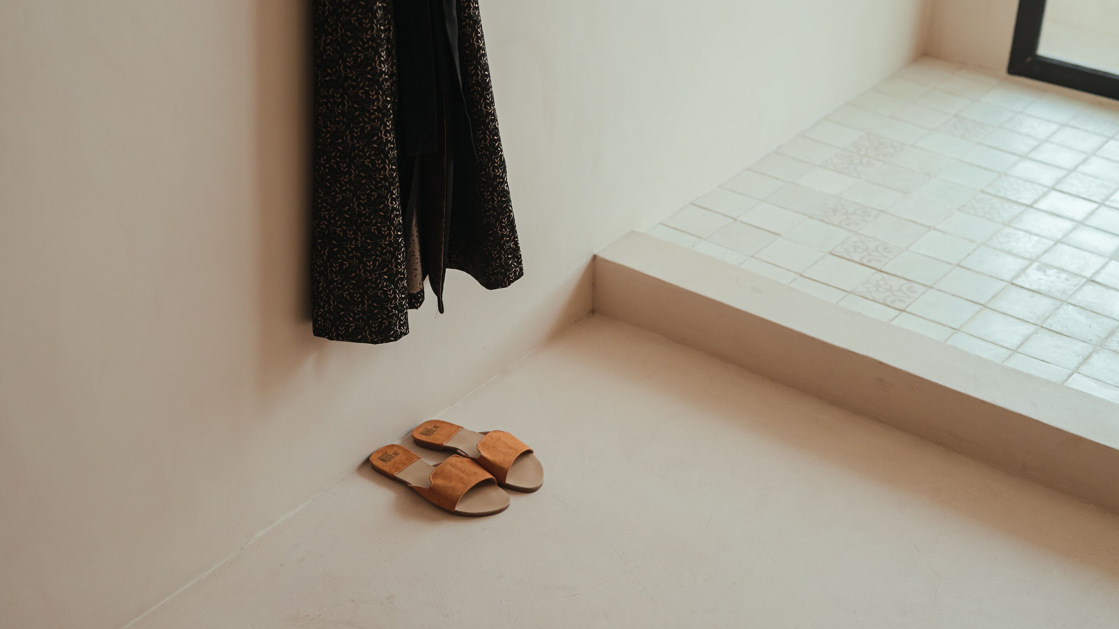 Un par de sandalias sobre un suelo de baldosas junto a una pared blanca con una ventana.